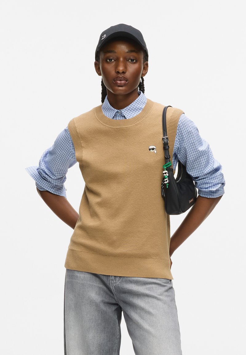 Kobieta z warkoczami, ubrana w beżowy sweter na koszulę w niebieską kratę, szare dżinsy, czapkę w kolorze czarnym i nosząca czarną torbę na ramię.