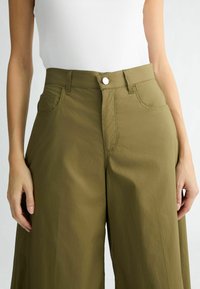 Pantalons larges en vert olive avec une texture lisse, fermeture par bouton et poches latérales, présentant des coutures détaillées sur le devant.