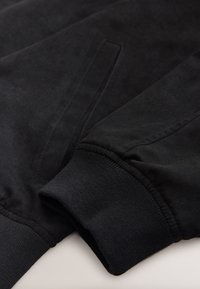 Gros plan sur la manche d'une veste noire montrant un poignet côtelé et une poche verticale fendue avec des bords cousus.