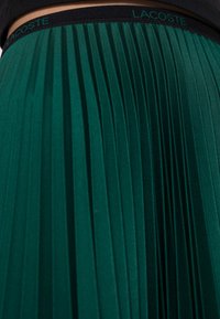 Vihreä pliseerattu hame, jossa on tekstuuripinta, mustalla vyötärönauhalla, johon on kohokuvioitu "LACOSTE". Pliseeraus luo dynaamisen visuaalisen vaikutelman.