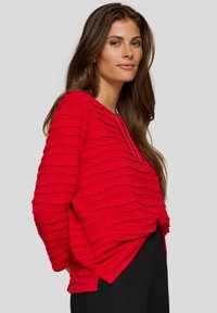 Frau mit langen braunen Haaren, die ein strukturiertes, leuchtend rotes Langarmoberteil und schwarze Hosen vor einem neutralen grauen Hintergrund trägt.