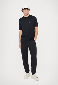 Črna majica s kratkimi rokavi z majhnim logotipom na prsih, kombinirana s temnimi športnimi hlačami in belimi supergami. Čist, minimalističen dizajn.