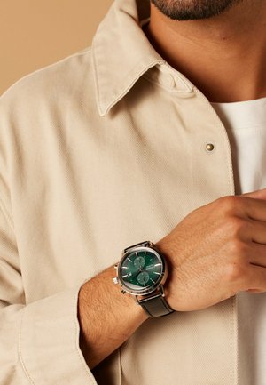 Ronde chronograafhorloge met een groene wijzerplaat, zilverkleurige accenten en een gestructureerde bruine leren band, gedragen om de pols met een beige overhemd.