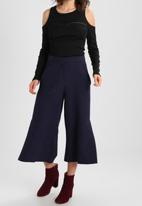 Black cold-shoulder top with textured accents, paired with wide-leg navy trousers. Model wears burgundy heeled ankle boots.