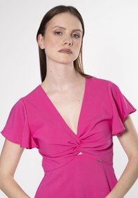 Fuchsia kjole med korte flutterærmer, dyb V-hals og snoet frontdetalje. Har en lille metallisk accent på siden.