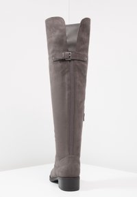 Bota de ante gris, por encima de la rodilla, con un panel trasero liso, cremallera lateral y un pequeño tacón cuadrado. Cuenta con una correa decorativa y detalle de hebilla.