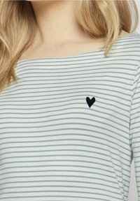 Gestreiftes Oberteil in Hellblau und Weiß, aus weichem Stoff. Hat einen weiten Ausschnitt und eine kleine schwarze Herz-Stickerei auf der Brust.
