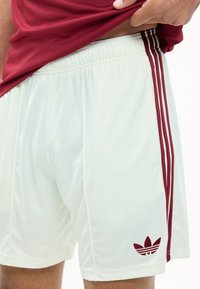 Λευκά αθλητικά σορτς με τρεις μαροον ρίγες στην πλευρά και λογότυπο Adidas σε μαροον κοντά στο τελείωμα, φορεμένα με ένα κόκκινο πουκάμισο που έχει ελαφρώς σηκωθεί.