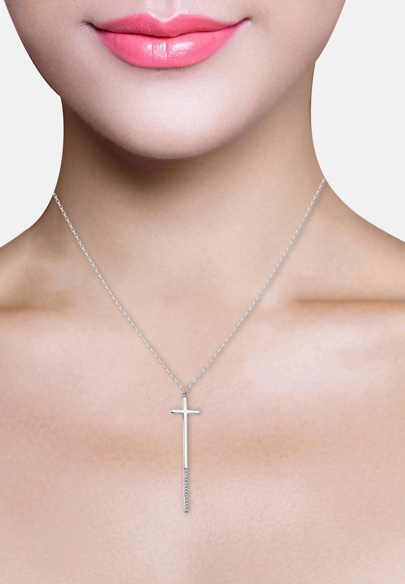 Srebrny krzyżyk na cienkim łańcuszku, o eleganckim, minimalistycznym wzornictwie. Delikatnie wisi nad gładkim, nieozdobionym dekoltem.