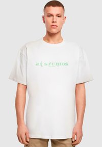 Hvid oversized T-shirt i bomuld med korte ærmer, der pryder teksten "21 STUDIOS EST MIX" i grønt på forsiden, simpel og minimalistisk design.