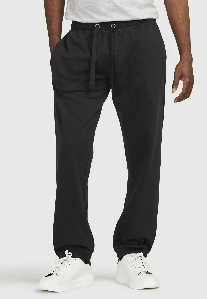 Sorte sweatpants med elastisk talje og snøre, der har sidelommer og elastiske manchetter ved anklerne. Bæres med hvide sneakers.