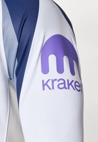 Hvid atletisk trøje med et lilla "kraken" logo på ærmet, med mørkeblå accenter og et tekstureret stofdesign.