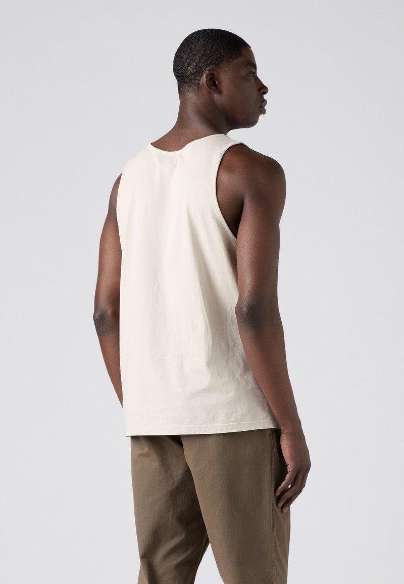 Homme portant un débardeur beige sans manches et un pantalon marron, se tenant de profil contre un fond gris clair uni.