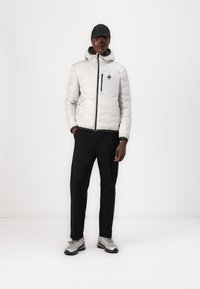 Giacca trapuntata grigia chiara con cappuccio, zip e tasca frontale; abbinata a pantaloni neri e scarpe sportive argentate.