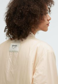 Hellbeige Bomberjacke mit glatter Textur, geripptem Kragen und einem Etikett mit der Aufschrift "SENSES the label" auf der Rückseite. Einfaches Design.