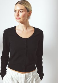 Črn prilagojen kardigan z okroglim izrezom, dolgimi rokavi in gumbom za zapiranje, v kombinaciji z belimi hlačami s tankimi črtami. Gladka tekstura.