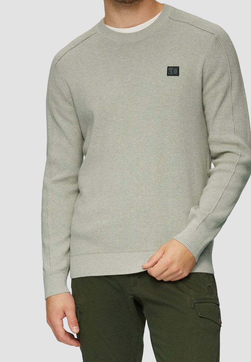 Homme portant un pull en tricot gris clair avec un petit logo noir sur la poitrine et un pantalon cargo vert foncé, debout devant un fond uni.