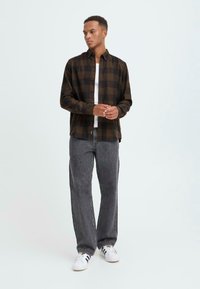 Bruine en zwarte geblokte flanellen shirt, knopen dicht, gedragen over een witte T-shirt; gecombineerd met grijze spijkerbroek en witte sneakers.