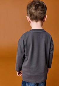 Graues Langarmshirt aus Baumwolle mit einfachem Design, getragen zu blauen Jeans, von hinten vor braunem Hintergrund gesehen.