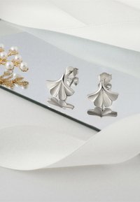 Silverörhängen formade som blommor med lager på lager av kronblad, visade på en reflekterande yta bredvid pärlor och mjukt vitt tyg.