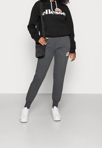 Schwarzer Hoodie mit weißem "ellesse"-Logo, kombiniert mit grauen Sweatpants, die ein kleines, farbiges Logo haben, und weißen Sneakers. Verstellbare Kapuze.