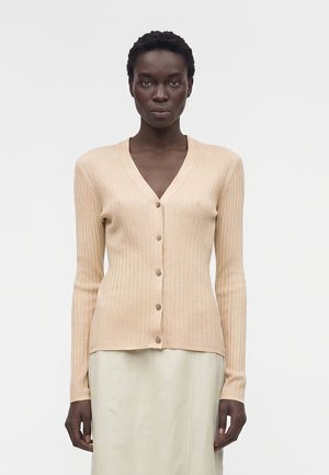 Žena s krátkymi kučeravými čiernymi vlasmi, oblečená v béžovom rebrovanom zapínanom kardigane a svetlej sukni, stojaca pred jednoduchým pozadím.