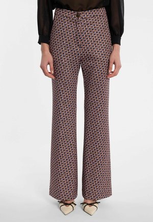 Femme portant un pantalon taille haute, jambes larges, avec un motif géométrique violet, marron et noir, associé à des chaussures pointues crème avec des brides noires.