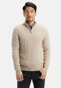 Pull en maille torsadée beige avec un col zippé, des poignets côtelés et un ourlet côtelé. Porté par-dessus une chemise sombre, présenté sur un mannequin masculin.