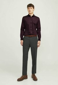 Tumši bordo krāsas krekls ar pogām un garām piedurknēm, kopā ar pelēkiem biksēm un brūnām ādas kurpēm, stāvot pret neitrālu fonu.