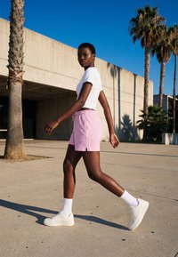 Jeune femme marchant en extérieur sur un trottoir en béton près de palmiers, portant des baskets blanches, des chaussettes, un short rose et un t-shirt court blanc.