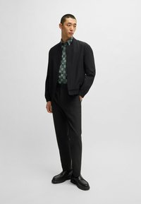 Giubbotto bomber nero sopra una camicia verde con motivo, pantaloni neri e scarpe nere. La camicia presenta un design con logo ripetuto.