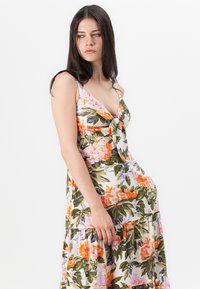 Blommig sommarklänning i krämfärg med orange, rosa och gröna mönster. Har en knytning fram, V-ringning och ett flerädlat kjoldesign med tunna band.