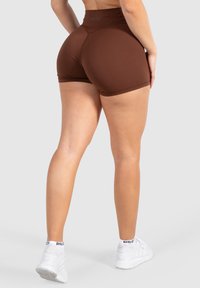 Bruine sportieve shorts met een hoge taille, gemaakt van rekbaar materiaal, met een naadloos ontwerp en een gladde textuur. Draagt een paar witte schoenen.