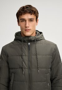 Giacca imbottita verde oliva con chiusura a zip, cappuccio regolabile e tasche frontali. Presenta una texture trapuntata e dettagli con cordoncino.