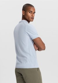 Lichtblauw gebreid poloshirt met korte mouwen, met ribbelstructuur aan de zijkanten en een standaardkraag. Gecombineerd met groene broek.