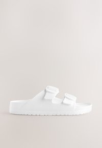 Sandales blanches à enfiler en caoutchouc, dotées de deux sangles ajustables. Texture lisse avec une semelle intérieure plate et profilée, ainsi qu'une semelle extérieure texturée.