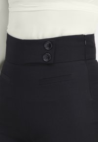 Pantalones negros de talle alto con dos botones delanteros, cremallera lateral y bolsillos con abertura, combinados con una camiseta blanca.