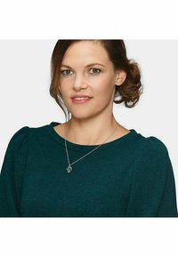 Silberne Halskette mit einem kreisförmigen Anhänger, der grüne Akzente aufweist. Das Modell trägt einen strukturierten türkisfarbenen Pullover mit Puffärmeln.