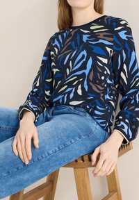 Personne assise sur un tabouret en bois portant un haut à manches longues à motif camouflage bleu et un jean denim bleu clair, les mains reposant sur les jambes.