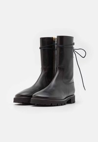 Bottes courtes en cuir noir à finition lisse, avec une fermeture éclair latérale et une semelle en caoutchouc texturée. Dotées de lacets noués autour de la cheville.