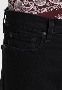 Czarne dżinsy z denimu o teksturowanej tkaninie, pięcioma kieszeniami i czarnymi metalowymi akcesoriami. Pas jest częściowo widoczny z detalami.