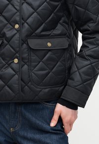 Μαύρο μπουφάν με καπιτονέ ύφασμα, με χρυσά πατζούρια και τσέπη με καπάκι, φορεμένο πάνω από σκούρο μπλε τζιν σε κορμό και χέρι ατόμου.