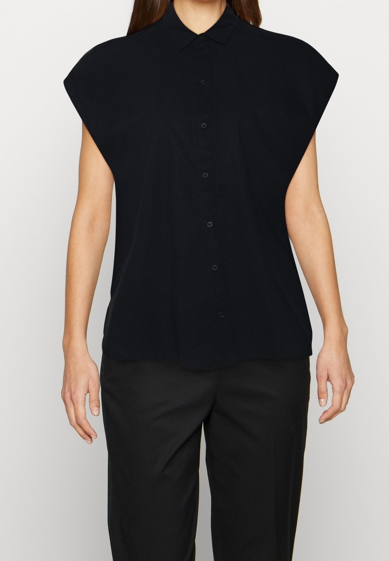 Camicia nera a maniche corte con colletto, con ampie spalle e una texture di tessuto liscia. Abbinata a pantaloni neri.