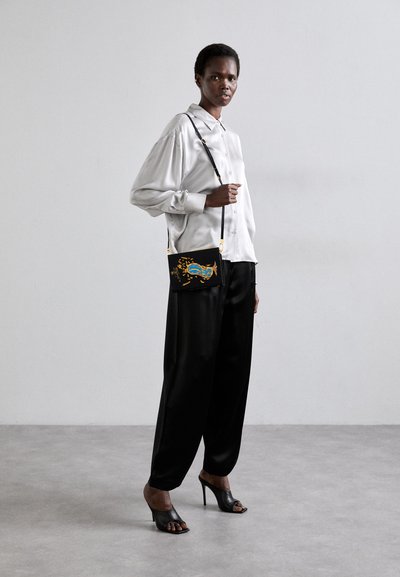 Sac à bandoulière noir avec des accents dorés présentant des motifs colorés et tourbillonnants ; modèle portant une chemise en soie blanche et un pantalon large noir.