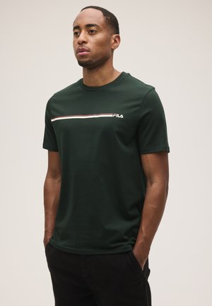 Uomo con capelli corti che indossa una t-shirt FILA verde scuro con una striscia bianca e rossa sul petto e pantaloni neri, mani nelle tasche.