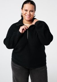 Zwarte pullover met ritssluiting in een gestructureerde brei, met een hoge kraag en een losse pasvorm. Gecombineerd met donkergrijze spijkerbroek.