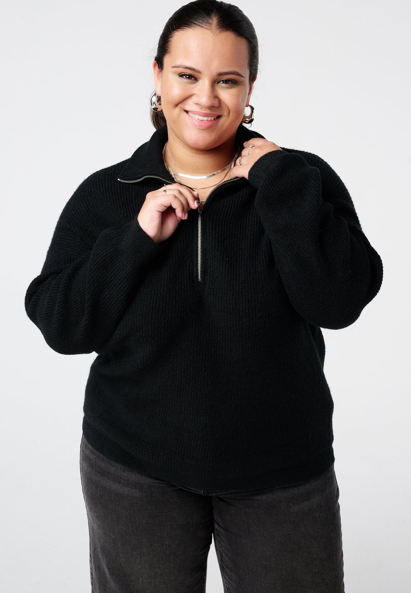 Zwarte pullover met ritssluiting in een gestructureerde brei, met een hoge kraag en een losse pasvorm. Gecombineerd met donkergrijze spijkerbroek.