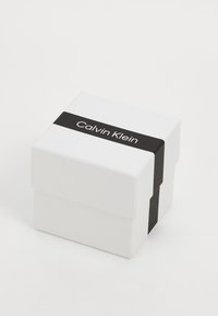 Baltas kartoninis dovanų dėžutė su juodu juostos, ant kurios užrašytas tekstas "Calvin Klein". Dėžutė turi paprastą, stačiakampį dizainą su lygia apdaila.