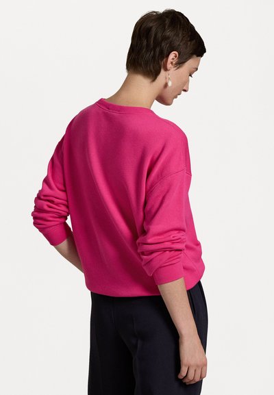 Vaaleanpunainen collegepaita, jossa on pyöreä kaula-aukko, ribattuja hihansuita ja rento istuvuus. Kangas näyttää pehmeältä ja sileältä.