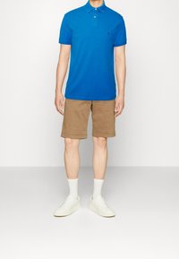 Blå piké i bomull, khakikortbyxor, vita sneakers, vita strumpor; avslappnad passform med knappslå och rullade ärmar.